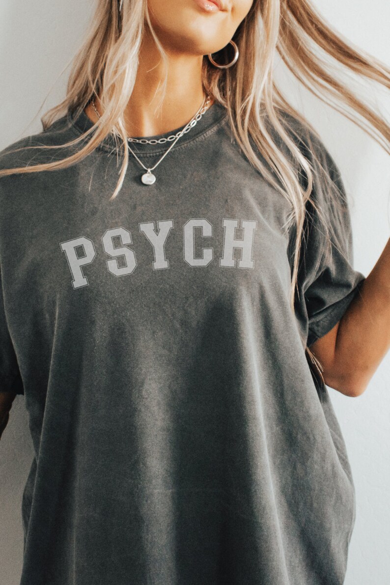 May include: A dark gray t-shirt with the word "PSYCH" printed in white on the front. The shirt is made of a soft, comfortable fabric and has a relaxed fit.