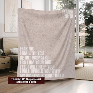 May include: A beige sherpa blanket with the text "ISAIAH 41:10" and a quote from the Bible. The blanket is displayed on a bed in a well-lit room. The text on the blanket reads "FEAR NOT, I AM WITH YOU..." and is in white.