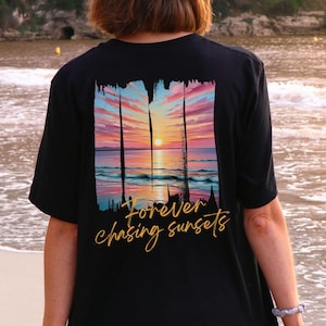 Może przedstawiać: Czarny t-shirt z grafiką zachodu słońca w odcieniach pomarańczu, r&oacute;żu i błękitu. Projekt zawiera napis "Forever chasing sunsets" w kursywie. Koszulka wykonana jest z miękkiego materiału.