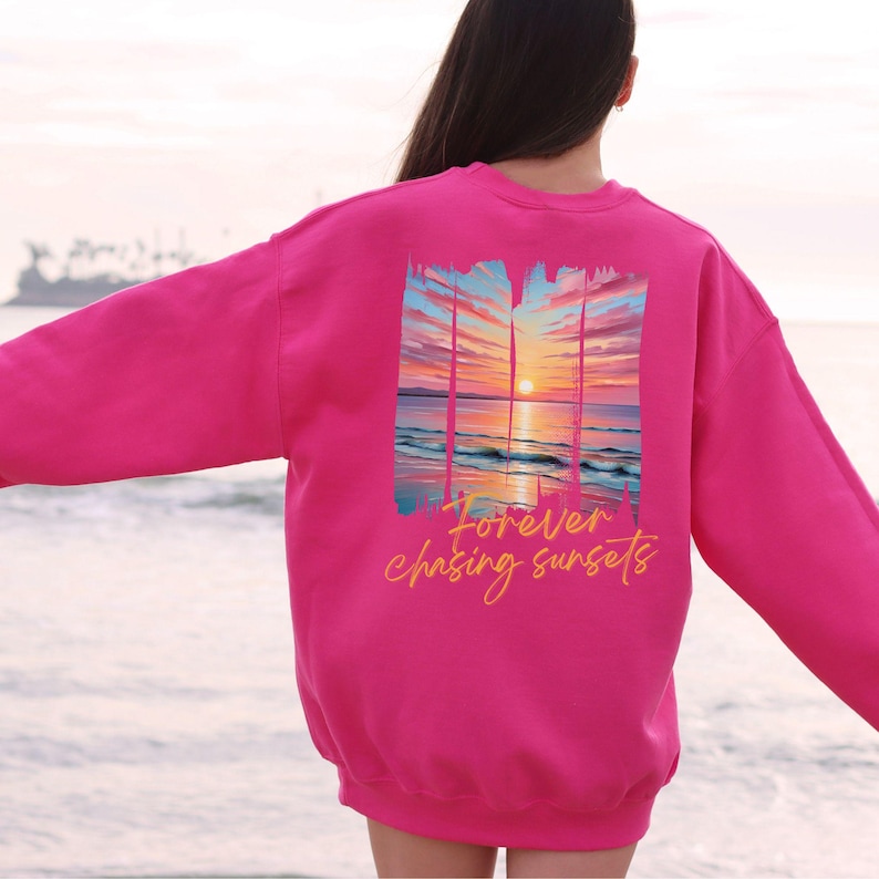 Może przedstawiać: R&oacute;żowa bluza z nadrukiem zachodu słońca na plecach. Grafika przedstawia żywy zach&oacute;d słońca nad oceanem z napisem "Forever chasing sunsets" w kursywie. Bluza jest w jednolitym kolorze.
