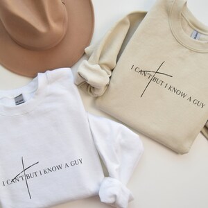 May include: Two sweatshirts with the text "I can't but I know a guy" and a stylized cross graphic. The sweatshirt on the left is white and the sweatshirt on the right is beige.