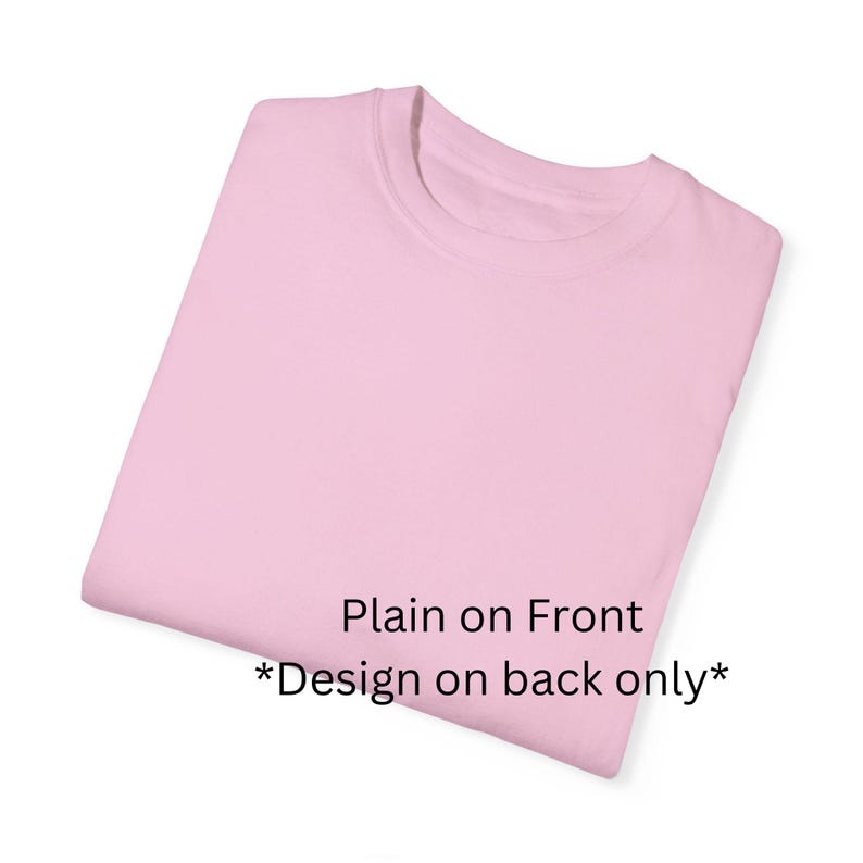 Może przedstawiać: Złożona, jasnor&oacute;żowa koszulka z okrągłym dekoltem. Prz&oacute;d koszulki jest gładki, z tekstem "Plain on Front *Design on back only*" wydrukowanym w prawym dolnym rogu.