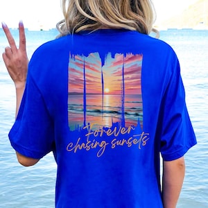 Może przedstawiać: Kr&oacute;lewsko niebieska koszulka z motywem zachodu słońca i złotym napisem "Forever chasing sunsets". Projekt zachodu słońca obejmuje żywe niebo, ocean i słońce.