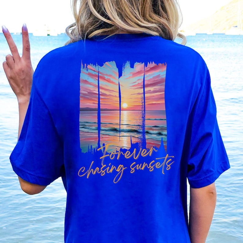 Może przedstawiać: Kr&oacute;lewsko niebieska koszulka z motywem zachodu słońca i złotym napisem "Forever chasing sunsets". Projekt zachodu słońca obejmuje żywe niebo, ocean i słońce.