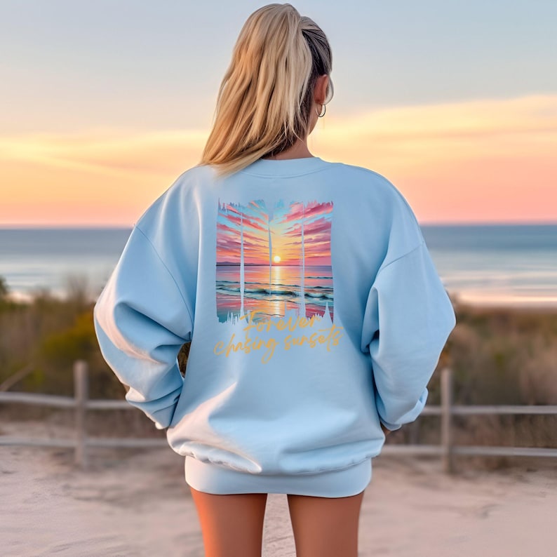 Może przedstawiać: Jasnoniebieska bluza z nadrukiem zachodu słońca na plecach. Grafika przedstawia kolorowy zach&oacute;d słońca nad oceanem z napisem "chasing sunsets" w ozdobnej czcionce. Bluza ma swobodny styl.