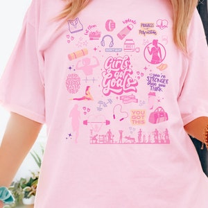 May include: Pink t-shirt with a graphic design of fitness-related icons and the text "Girls got Goals". The design includes images of weights, a yoga mat, a jump rope, a protein shake, and a gym bag.