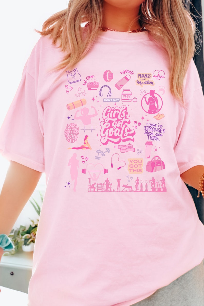 May include: Pink t-shirt with a graphic design of fitness-related icons and the text "Girls got Goals". The design includes images of weights, a yoga mat, a jump rope, a protein shake, and a gym bag.