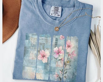 May include: A blue Comfort Colors t-shirt with a floral design. The graphic features pink flowers and green leaves against a blue background. A gold necklace with a pendant rests on the shirt. The shirt is folded.