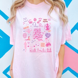 May include: Pink t-shirt with a graphic design of fitness and motivational quotes. The design includes images of weights, a yoga mat, a water bottle, and a heart with the text "Girls got goals".