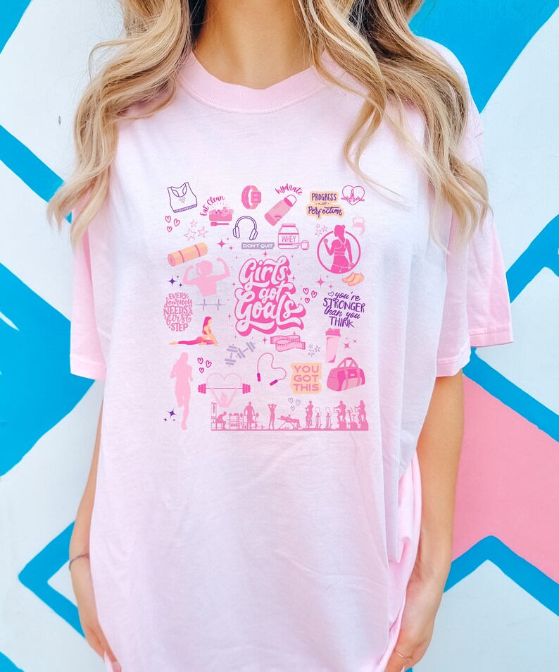 May include: Pink t-shirt with a graphic design of fitness and motivational quotes. The design includes images of weights, a yoga mat, a water bottle, and a heart with the text "Girls got goals".