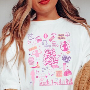 May include: White t-shirt with a pink and purple design featuring the text "Girls got Goals" and various fitness-related icons, including a heart, dumbbells, a yoga mat, a water bottle, and a jump rope.
