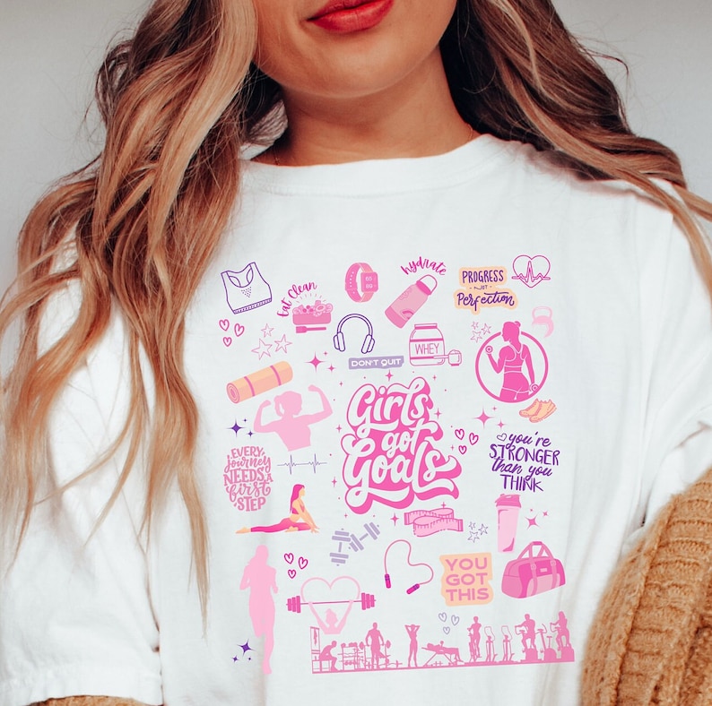 May include: White t-shirt with a pink and purple design featuring the text "Girls got Goals" and various fitness-related icons, including a heart, dumbbells, a yoga mat, a water bottle, and a jump rope.