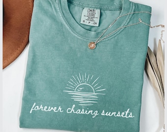 May include: A teal Comfort Colors t-shirt with the phrase "forever chasing sunsets" printed in white. The design includes a sun setting over the ocean. A silver necklace with a pendant is draped across the shirt.
