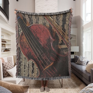 May include: A woven throw blanket featuring a detailed illustration of a violin in shades of red, brown, and black. The design includes the instrument's strings and bow against a dark background with a fringe border.