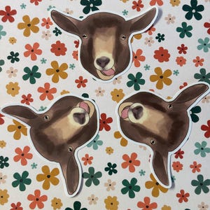 May include: Three brown and white goat head stickers with a floral background. The goats are looking at the camera with their tongues sticking out.