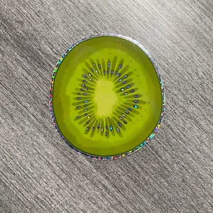 May include: A round sticker with a holographic finish featuring a green kiwi fruit slice. The kiwi has a brown seed pattern and a light green center.