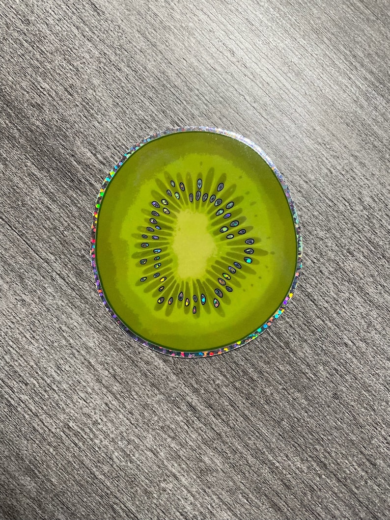 Może przedstawiać: Okrągła naklejka z holograficznym wykończeniem przedstawiająca plaster zielonego kiwi. Kiwi ma brązowy wz&oacute;r nasion i jasnozielone centrum.