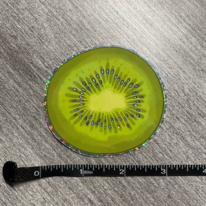 Może przedstawiać: Holograficzna naklejka przedstawiająca plaster kiwano. Kiwi jest zielone z brązowym środkiem i czarnymi pestkami. Naklejka ma średnicę około 5 cm.