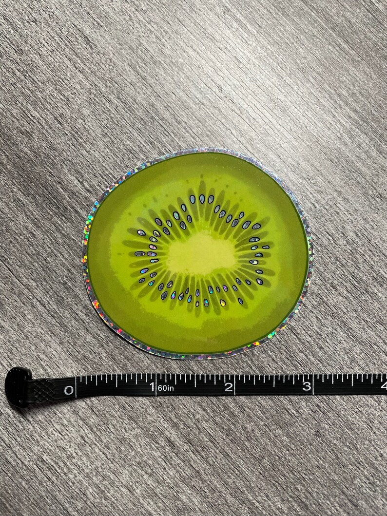 Może przedstawiać: Holograficzna naklejka przedstawiająca plaster kiwano. Kiwi jest zielone z brązowym środkiem i czarnymi pestkami. Naklejka ma średnicę około 5 cm.