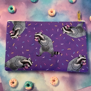 May include: Purple zippered pouch with a repeating pattern of raccoons eating donuts and colorful sprinkles on a white background.