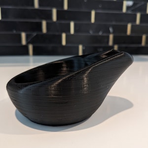 May include: A black 3D printed bowl with a smooth, rounded base and a distinctively angled, elevated rim. The surface features visible horizontal layer lines characteristic of the additive manufacturing process, creating a textured finish.