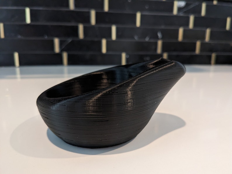 May include: A black 3D printed bowl with a smooth, rounded base and a distinctively angled, elevated rim. The surface features visible horizontal layer lines characteristic of the additive manufacturing process, creating a textured finish.