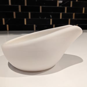 May include: A smooth, white ceramic bowl with a curved, bird-like shape. The bowl has a wide opening and a rounded base. The design is minimalist and modern, suitable for serving or display. The bowl is approximately 20 cm long.