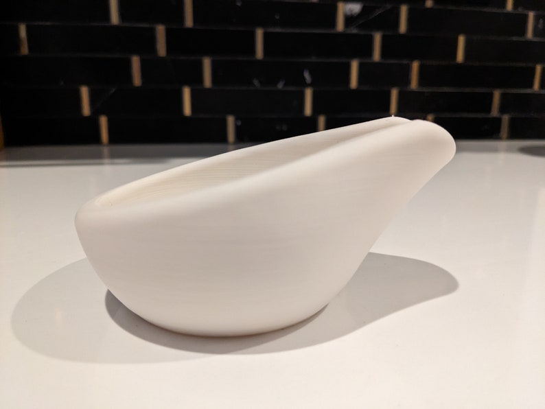 May include: A smooth, white ceramic bowl with a curved, bird-like shape. The bowl has a wide opening and a rounded base. The design is minimalist and modern, suitable for serving or display. The bowl is approximately 20 cm long.