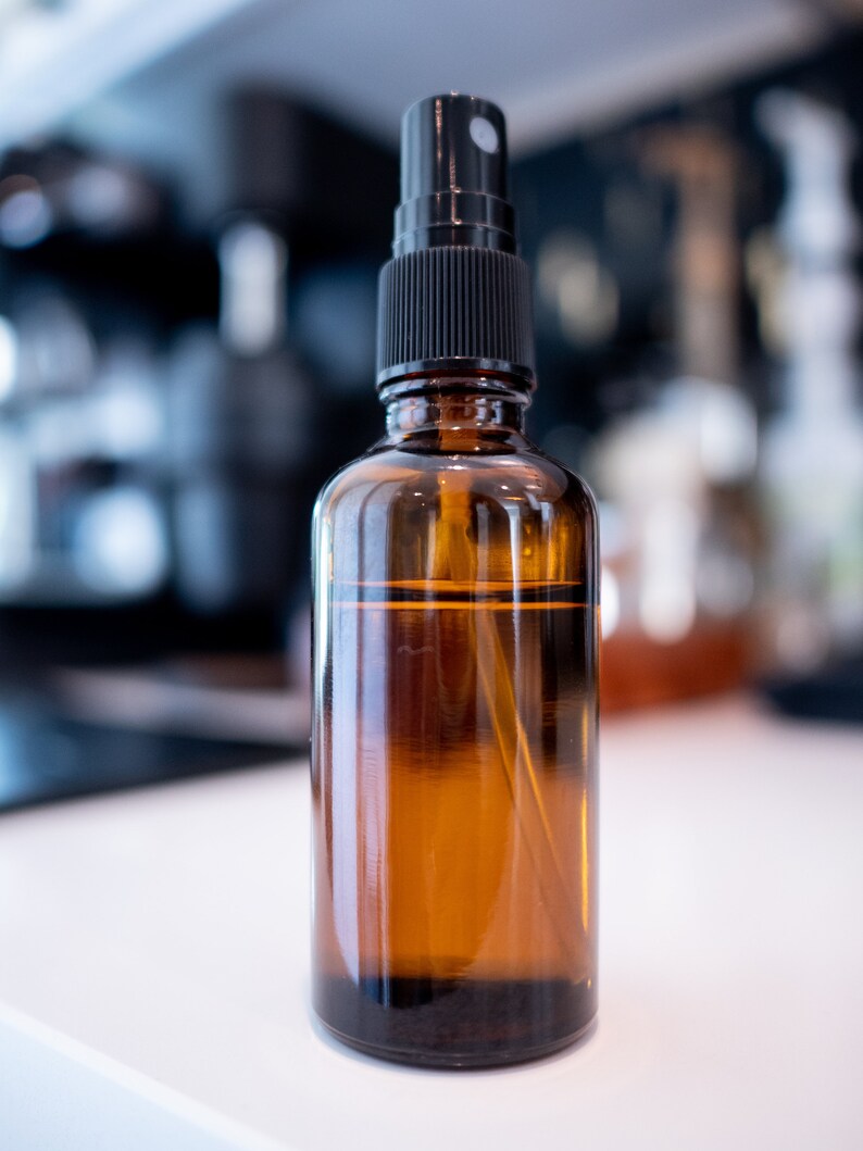 May include: An amber glass spray bottle with a black spray nozzle. The bottle is cylindrical and contains a clear liquid. The background is blurred, with other objects visible.