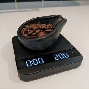 May include: A black digital kitchen scale shows "0:00" and "20.0" with a small black bowl filled with roasted coffee beans. The bowl is dark grey and has a unique shape. The scale is on a white surface.