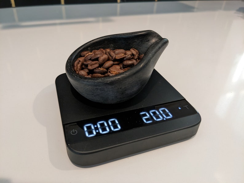 May include: A black digital kitchen scale shows "0:00" and "20.0" with a small black bowl filled with roasted coffee beans. The bowl is dark grey and has a unique shape. The scale is on a white surface.