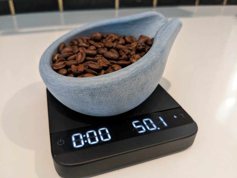 May include: A light blue, textured ceramic scoop filled with roasted coffee beans, resting on a black digital scale. The scale displays a weight of 1422 grams.