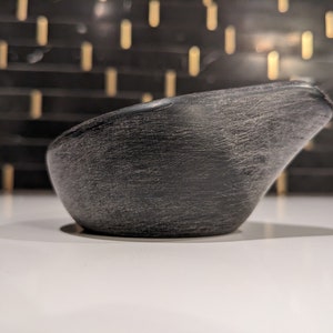 May include: A dark grey, textured bowl with a spout. The bowl has a smooth, curved shape and a rough, weathered surface. The background is a blurred pattern of black and gold.