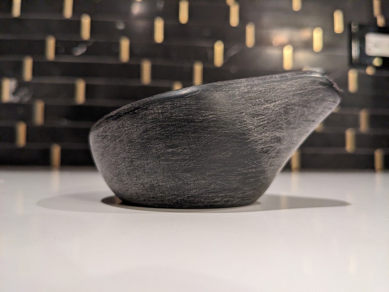 May include: A dark grey, textured bowl with a spout. The bowl has a smooth, curved shape and a rough, weathered surface. The background is a blurred pattern of black and gold.