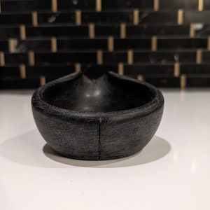 May include: A small, black, wooden bowl with a distinctive, curved form. The bowl has a textured surface and a smooth interior. The design includes a small indentation on one side. The bowl is likely for serving or display.