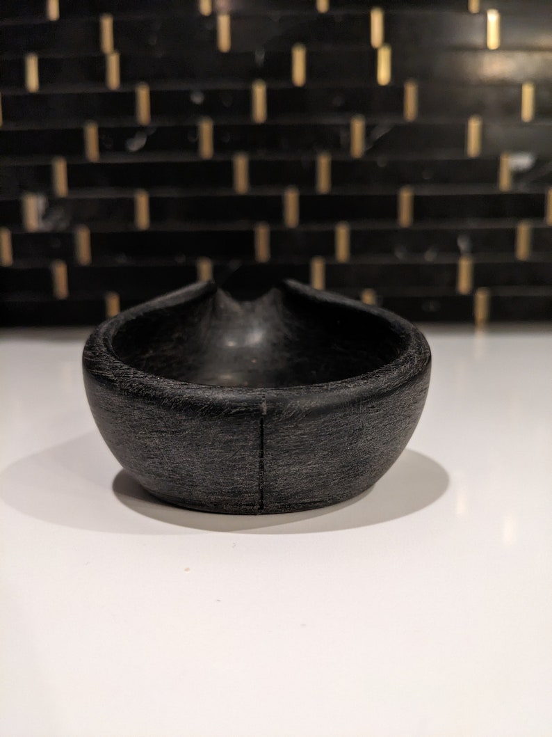 May include: A small, black, wooden bowl with a distinctive, curved form. The bowl has a textured surface and a smooth interior. The design includes a small indentation on one side. The bowl is likely for serving or display.