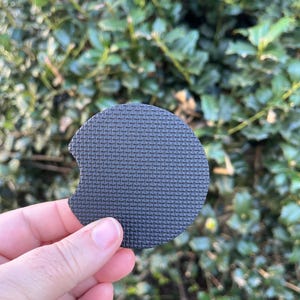 May include: A black round coaster with a textured surface. The coaster has a cutout on one side.