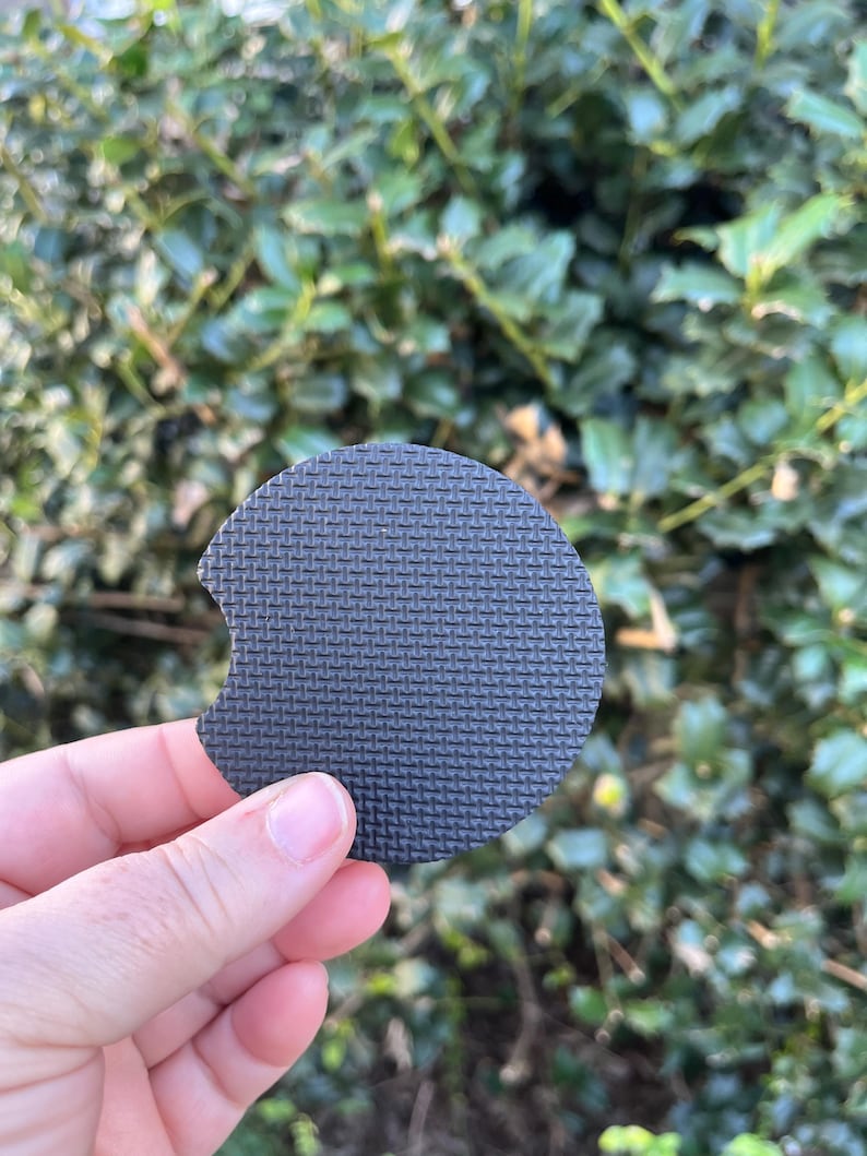 May include: A black round coaster with a textured surface. The coaster has a cutout on one side.