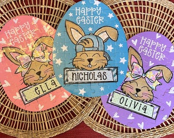 May include: Three personalized Easter egg puzzles with bunny illustrations. The blue puzzle says "Happy Easter Nicholas", the pink puzzle says "Happy Easter Ella", and the purple puzzle says "Happy Easter Olivia".
