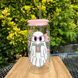 May include: A clear glass reusable water bottle with a pink lid and a straw. The bottle has a graphic of a ghost wearing glasses and reading a book. The ghost is surrounded by pink flowers.