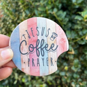 May include: A round, colorful coaster with a wood grain pattern. It features the text "Jesus Coffee Prayer" and a small coffee cup icon.