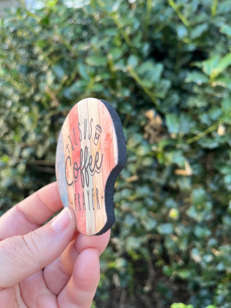 May include: A round wooden coaster with a distressed woodgrain design. The coaster features the text "Jesus + Coffee = Prayer" in black lettering with a coffee cup icon.