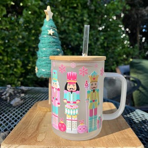 May include: A frosted glass mug with a bamboo lid and a clear straw. The mug features a colorful design of nutcracker figures and snowflakes. A small green Christmas tree is in the background, adding to the festive theme.