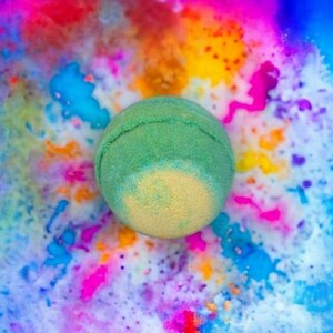 May include: A green bath bomb with a yellow centre sits against a vibrant background of colourful splashes. The bath bomb is round and textured, with a visible seam. The background features splashes of pink, blue, orange, and yellow.
