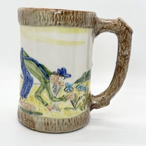 May include: A ceramic mug with a whimsical design. The mug features a brown tree-branch handle and a painted scene of a figure in a blue hat and green outfit tending to blue flowers. The mug has a light brown rim and base.