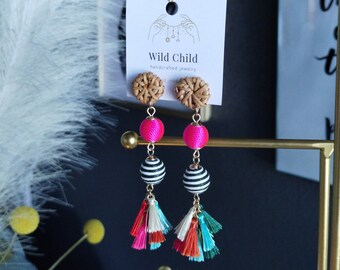 May include: A pair of handmade earrings with a woven wood top, a pink and white striped ball, and a multi-coloured tassel. The earrings are displayed on a gold stand with a sign that reads "Wild Child handcrafted jewellery".