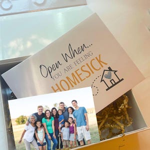 K&ouml;nnte beinhalten: Eine wei&szlig;e Geschenkbox mit goldenem F&uuml;llpapier und einem Foto einer zehnk&ouml;pfigen Familie. Der Deckel tr&auml;gt den schwarz-goldenen Text "Open When... YOU ARE FEELING HOMESICK" sowie ein Haus-Symbol mit einem Herz und einer gestrichelten Linie.