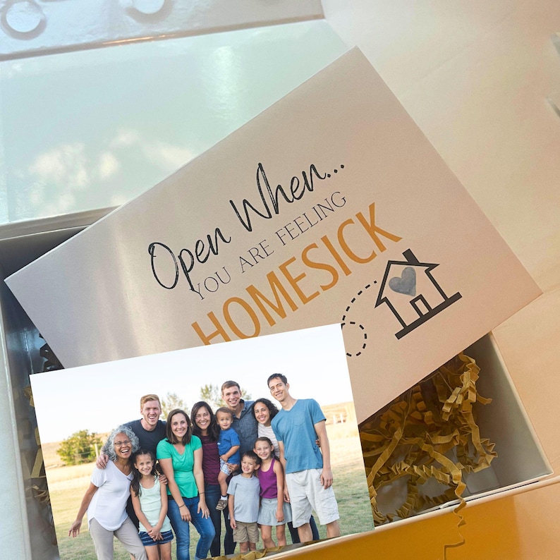 K&ouml;nnte beinhalten: Eine wei&szlig;e Geschenkbox mit goldenem F&uuml;llpapier und einem Foto einer zehnk&ouml;pfigen Familie. Der Deckel tr&auml;gt den schwarz-goldenen Text "Open When... YOU ARE FEELING HOMESICK" sowie ein Haus-Symbol mit einem Herz und einer gestrichelten Linie.
