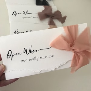 May include: A set of white cards with the text "Open When" followed by a phrase. The cards are tied with pink and brown ribbon.