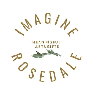 May include: Gold text on a white background reads "Imagine Rosedale" in a circular shape. The text "Meaningful Art & Gifts" is centred below the circle. A sprig of green eucalyptus leaves is centred below the text.